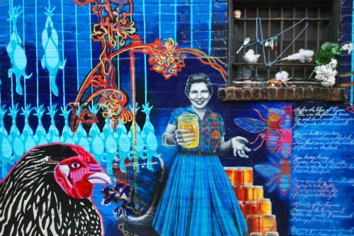Colorful mural featuring a woman in a blue dress holding a jar, surrounded by vibrant patterns and a chicken.