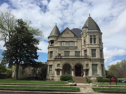 A grand, historic stone mansion with turrets and intricate architectural details, surrounded by greenery and a blue sky.