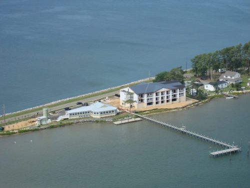 Aerial view of a waterfront property with a large building, dock, and surrounding greenery by the water.