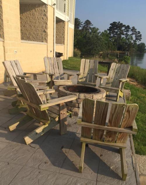 A circular arrangement of wooden chairs around a stone fire pit by a calm lake, with trees in the background.