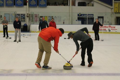 Two curlers compete on the ice, sweeping a stone while others watch in the background at a curling rink.