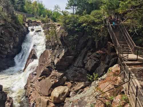 A waterfall cascades over rocky cliffs, with a wooden walkway and lush greenery surrounding the scene.