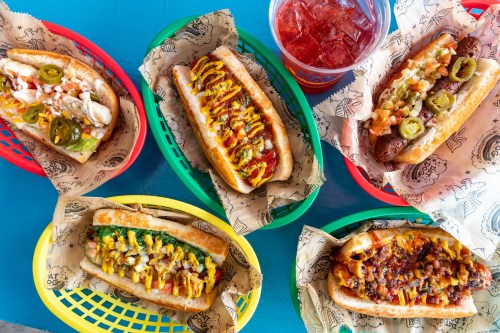 A colorful assortment of hot dogs with various toppings, served in baskets, alongside a drink.