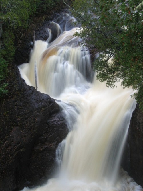 A cascading waterfall flows over rocky terrain, surrounded by lush greenery and trees.