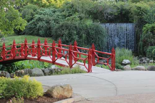 A red bridge arches over a path near a waterfall, surrounded by lush greenery and rocks.