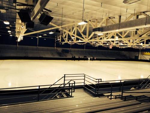 An empty ice rink with bleachers, illuminated by overhead lights, creating a quiet, serene atmosphere.