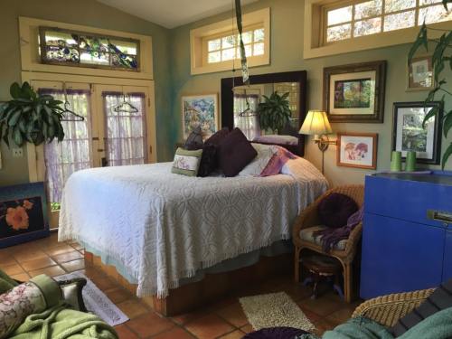 Cozy bedroom with a large bed, colorful decor, plants, and natural light from windows. Warm, inviting atmosphere.