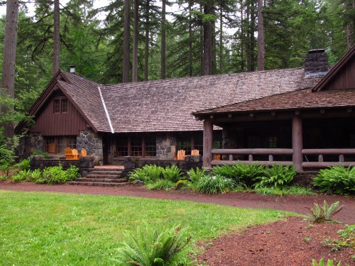 A rustic cabin surrounded by tall trees, featuring a stone facade and wooden porch with chairs. Lush greenery in the foreground.