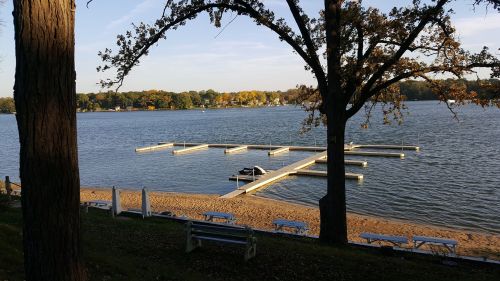 A serene lakeside view with a dock, sandy beach, and trees, showcasing autumn colors under a clear sky.