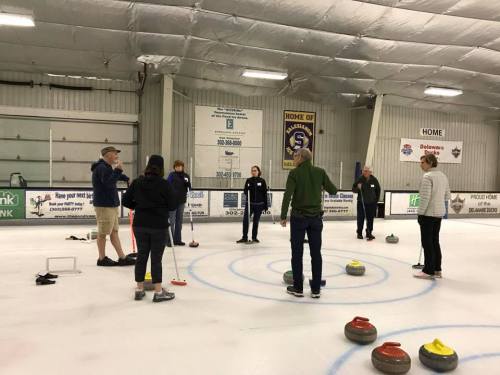 A group of people stands on an ice rink, discussing curling techniques with stones scattered around.