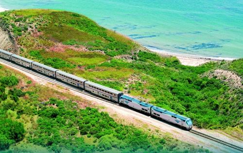 A train travels along a scenic coastal route, surrounded by lush greenery and blue waters.