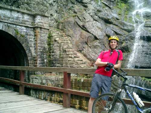 A cyclist in a red shirt and yellow helmet leans against a wooden railing near a stone tunnel and waterfall.