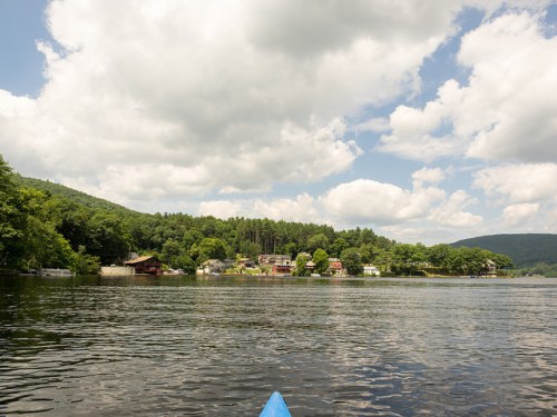 A serene lake scene with lush green trees, houses along the shore, and fluffy clouds in a blue sky.