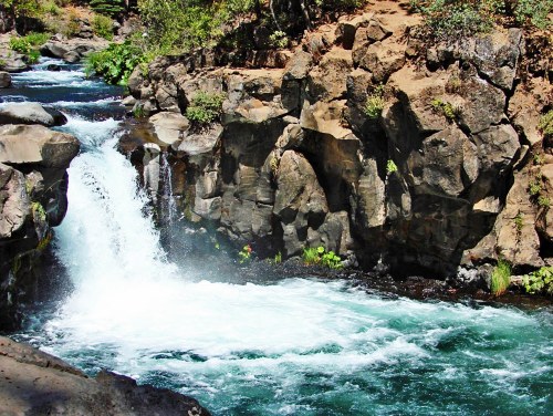 A serene waterfall cascades over rocky cliffs into a clear, turquoise river surrounded by lush greenery.
