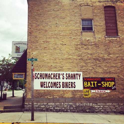 Sign for Schumacher's Shanty welcoming bikers, with a bait shop sign on a brick building in a small town.