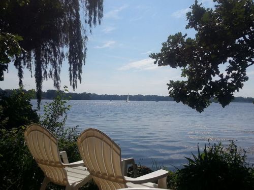 A serene lakeside view with two white chairs, lush greenery, and a sailboat in the distance under a clear blue sky.