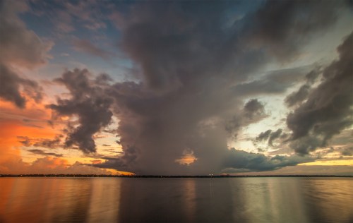 A dramatic sunset over a calm water body, featuring vibrant clouds and a looming storm in the distance.