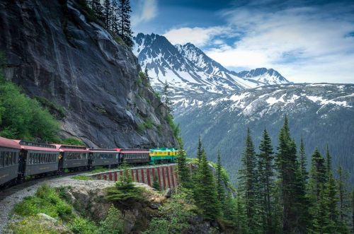 A colorful train travels along a scenic mountain route, surrounded by lush trees and snow-capped peaks.