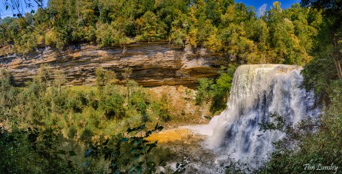 A panoramic view of a waterfall cascading over rocky cliffs, surrounded by lush green trees and a clear blue sky.