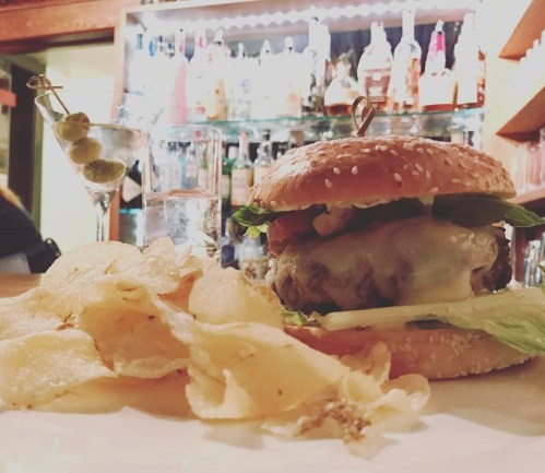 A juicy burger with lettuce, tomato, and cheese, served with potato chips and a martini in the background.