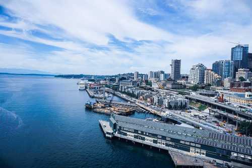 A scenic view of a waterfront city with buildings, docks, and a calm blue sea under a partly cloudy sky.