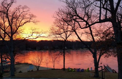 Sunset over a calm lake, with silhouetted trees and colorful reflections on the water.