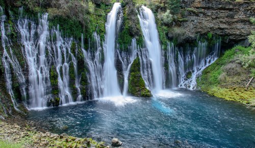 A serene waterfall cascading over rocky cliffs into a clear blue pool, surrounded by lush greenery.