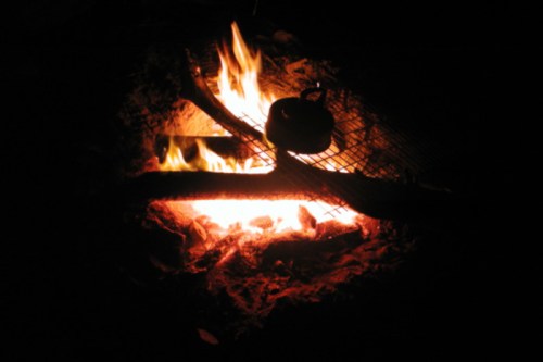 A campfire burns brightly at night, with a kettle hanging over the flames on a grill above.