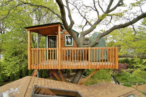 A cozy treehouse with a wooden deck, nestled among green leaves and branches, overlooking a garden below.