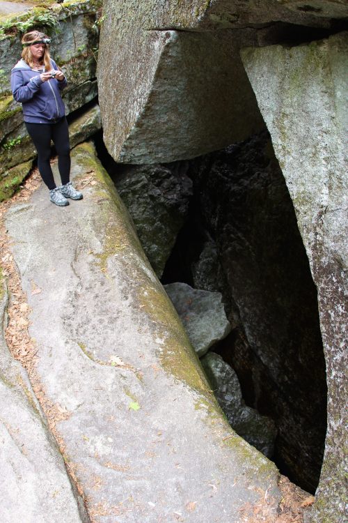 A person stands near a deep crevice between large rocks in a forested area, holding a phone and looking down.