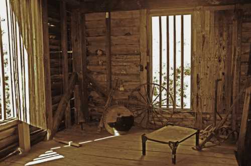 Interior of a rustic, wooden room with a table, wheel, and sunlight streaming through barred windows.