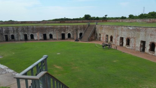 A historic fort with a grassy courtyard, brick walls, and cannons, surrounded by a staircase and open sky.