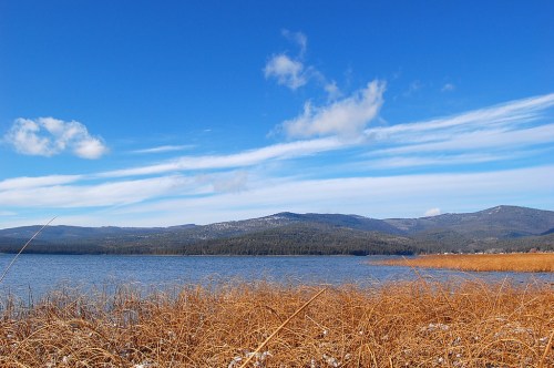 A serene lake surrounded by golden grasses and mountains under a clear blue sky with wispy clouds.