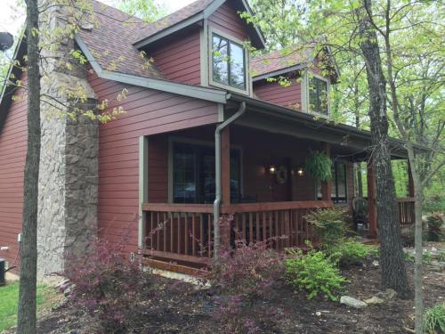 A cozy red house with a stone chimney, surrounded by trees and greenery, featuring a welcoming front porch.