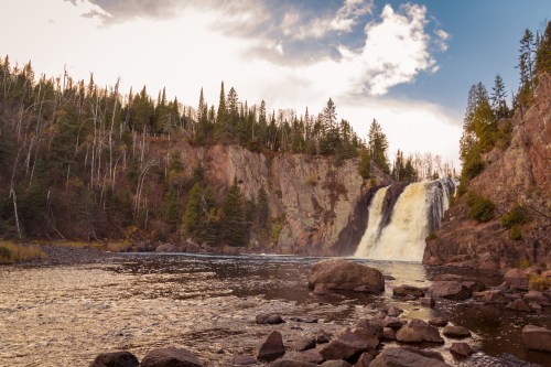 A scenic waterfall cascades into a river, surrounded by rocky shores and lush green trees under a cloudy sky.