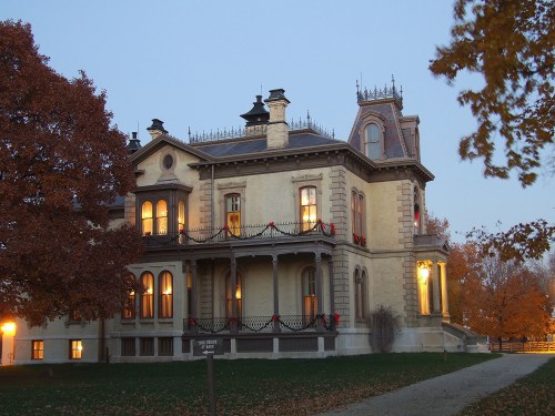 A historic mansion adorned with holiday decorations, surrounded by autumn trees at dusk. Warm lights glow from the windows.