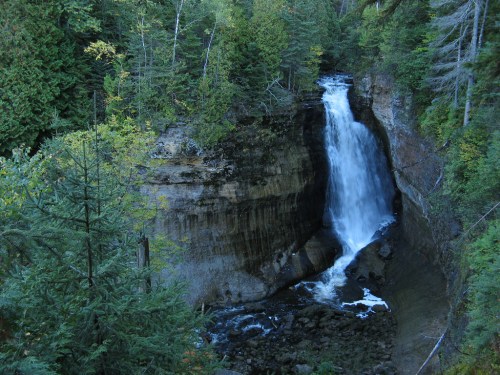 A serene waterfall cascades down rocky cliffs, surrounded by lush green trees and a tranquil forest setting.