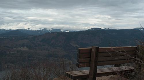 A wooden bench overlooks a mountainous landscape under a cloudy sky. Snow-capped peaks are visible in the distance.