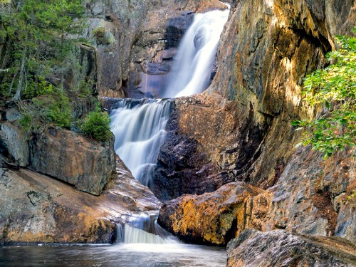 A serene waterfall cascading over rocky cliffs, surrounded by lush greenery and sunlight filtering through the trees.