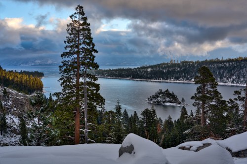 A serene winter landscape featuring a lake surrounded by snow-covered trees and distant mountains under a cloudy sky.