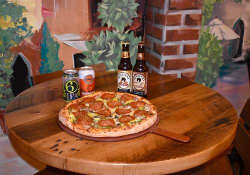 A wooden table with a pepperoni pizza and two bottles of beer, alongside a can of IPA, set against a colorful mural.