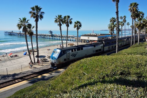 A train passes by a beach with palm trees, near a pier and people enjoying the sun on the sand.