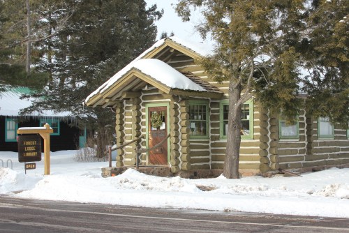 A log cabin library covered in snow, with a sign reading "Forney Library" in front. Trees are visible nearby.