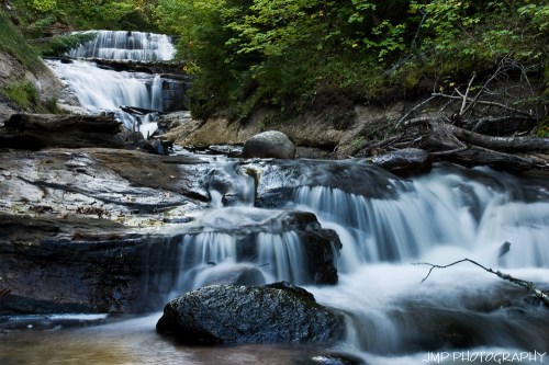A serene waterfall cascades over rocks, surrounded by lush greenery and a tranquil forest setting.