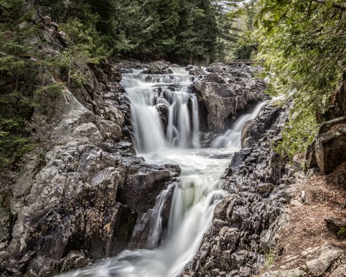 A serene waterfall cascades over rocky terrain, surrounded by lush green trees and a peaceful natural setting.