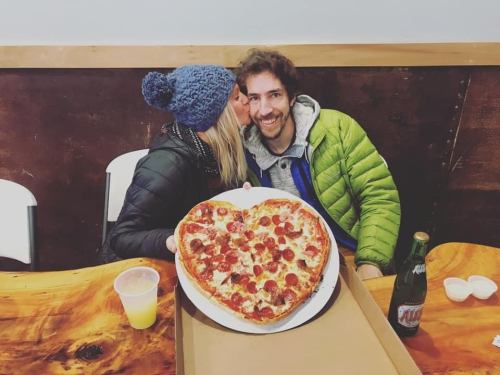 A couple shares a kiss beside a large heart-shaped pizza on a table, with drinks in the background.