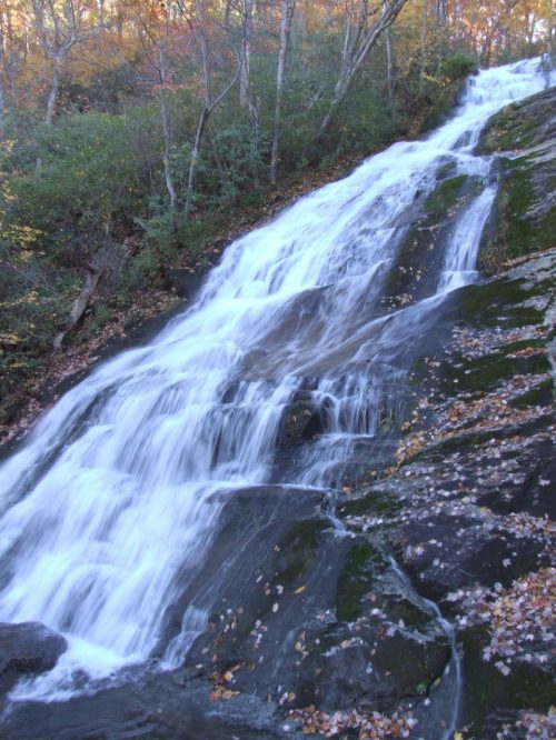 A cascading waterfall flows over rocky terrain, surrounded by autumn foliage and greenery.