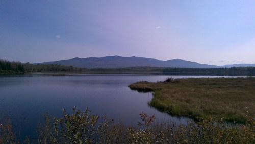 A serene lake surrounded by lush greenery and distant mountains under a clear blue sky.