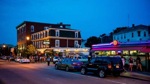 A lively street scene at dusk, featuring restaurants, cars parked along the road, and people waiting outside.