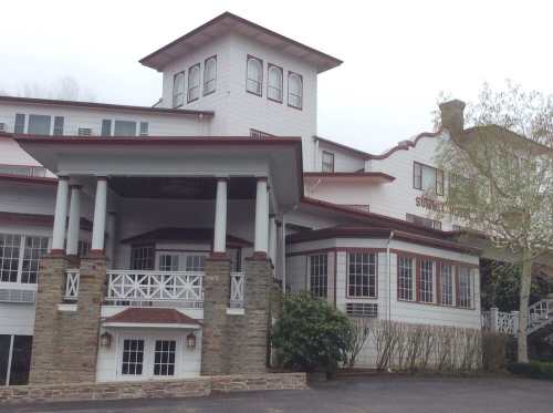 A large, multi-story white building with a red roof and decorative porch, surrounded by greenery.